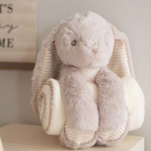 Light brown plush rabbit with cream corduroy ears holding a rolled cream blanket.